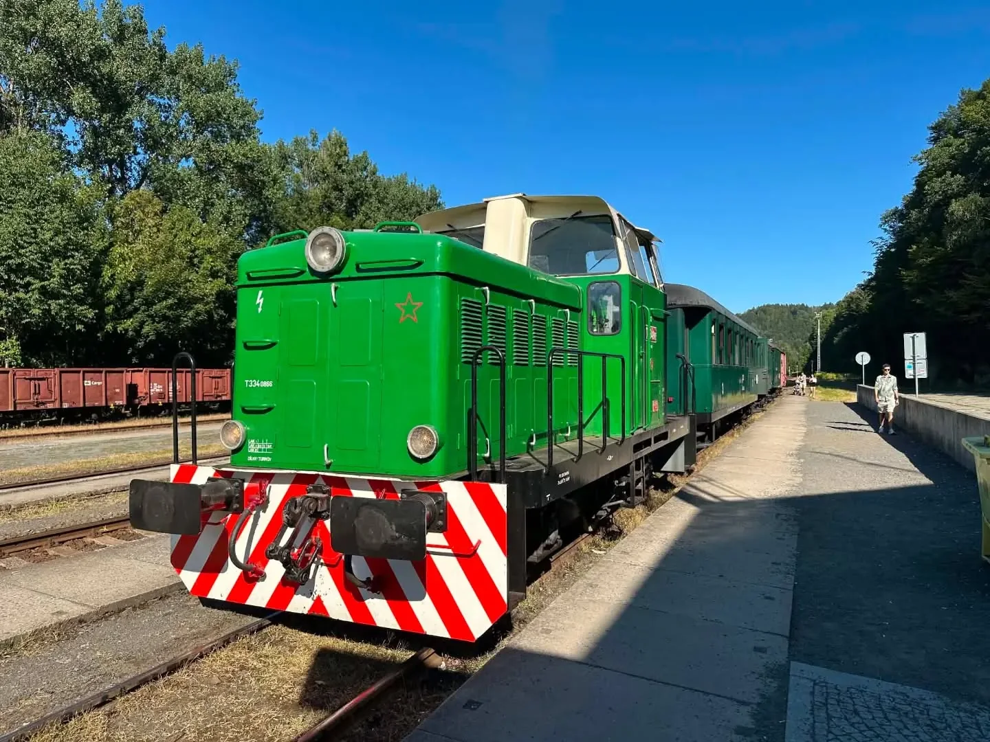 Zelená historická lokomotiva s osobními vozy stojí u nástupiště.